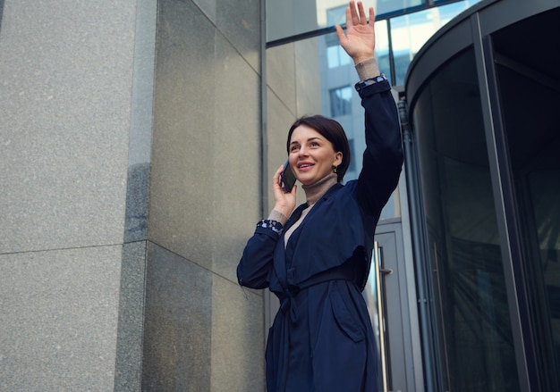 Self-confident beautiful woman smiling with toothy smile, talking on a mobile phone and raising her hand when meeting with someone while leaving a modern corporate high-rise building