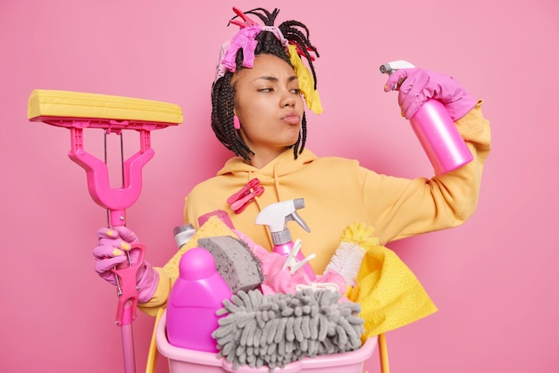 Self confident proud housewife holds cleaning detergent and mop for washing floor keeps flat tidy poses near basket with rags and cleaning supplies does domestic duties isolated on pink background