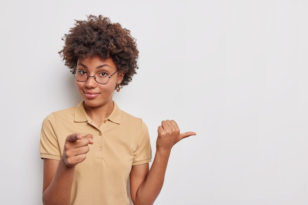 Self confident woman points at your and away on blank space confirms to select this wears round spectacles casual t shirt attracts attention