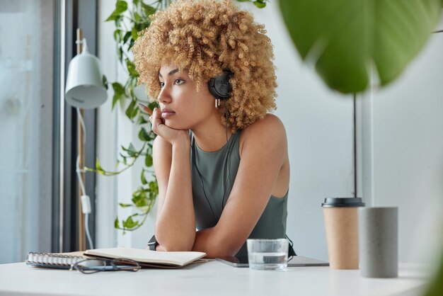 Self education distance learning concept thoughtful curly\
female model sits at workplace desk against cozy interior wears\
headset listens training webinar online drinks coffee focused in\
window