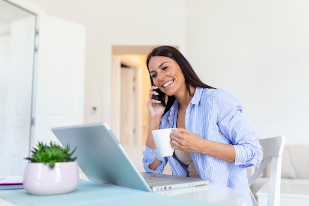 Self employed woman working with her phone and laptop at home\
with a cup of coffee happy woman using mobile phone while working\
at home with laptop smiling woman messaging with smartphone