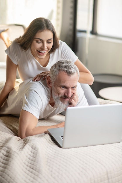 Self-employment. Bearded smiling man working on laptop at home while lying on bed and joyful woman near