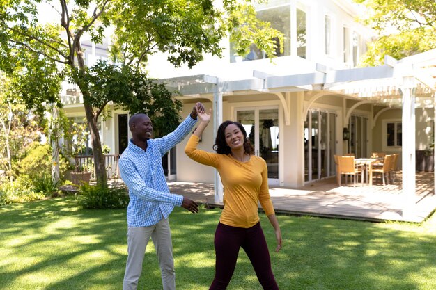 Photo self isolation in quarantine lock down. front view of an african american man and a mixed race woman outside their house in the garden on a sunny day, having fun holding hands and dancing together