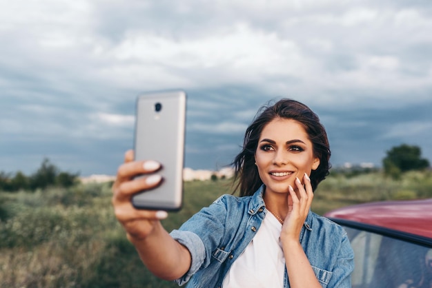 Self portrait of beautiful young brunette smile womantaking selfie on nature and cloudy sky background near to red car Travel lifestyle and Instagram concept
