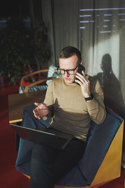 A selfemployed man in stylish clothes is making a blog website post on a laptop computer in a modern workspace Male freelancer works remotely and talks on the phone