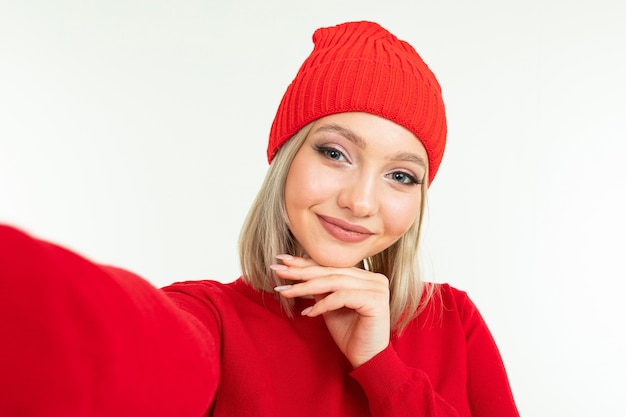 Selfie of a cute blonde girl in a red hat and sweater on a white background.