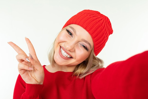 Selfie of a cute blonde girl in a red hat and sweater on a white background.