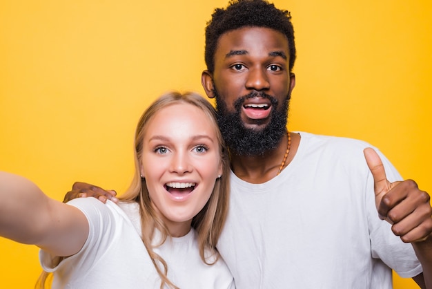 Selfie Fun. Cheerful interrational couple taking self-portrait together, looking at camera and smiling, posing over yellow wall