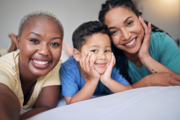 Selfie gay family and happy on a bed in home bedroom for security quality time and love Adoption lesbian or LGBTQ women or interracial parents and happy kid together for social media memory