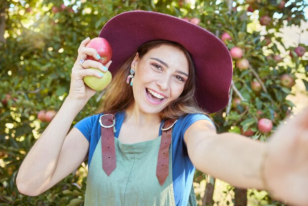 2つの異なるリンゴを持っている果樹園に立っている幸せな女性農家の自分撮り晴れた日に麦わら帽子とダンガリーを身に着けている1人の若い笑顔の農家の労働者の肖像画果物狩り