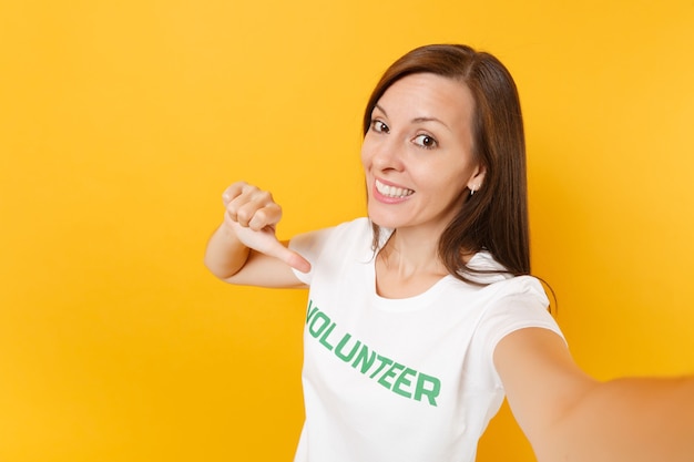 Selfie image of happy smiling satisfied woman in white t-shirt with written inscription green title volunteer isolated on yellow background. Voluntary free assistance help, charity grace work concept.