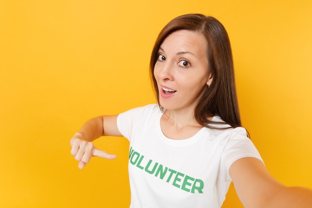 Selfie image of happy smiling satisfied woman in white t-shirt with written inscription green title volunteer isolated on yellow background. Voluntary free assistance help, charity grace work concept.