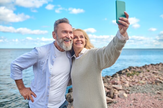 Selfie in de natuur. blonde vrouw met smartphone in haar uitgestrekte hand die overdag selfie maakt met lachende bebaarde man buiten