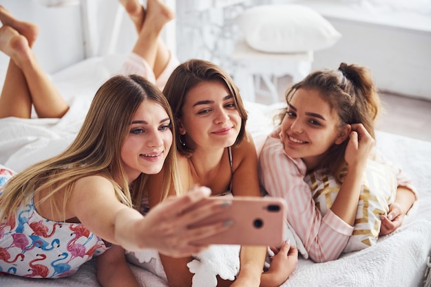 Selfie maken met behulp van roze telefoon. Gelukkige vrouwelijke vrienden die plezier hebben op een pyjamafeestje in de slaapkamer.