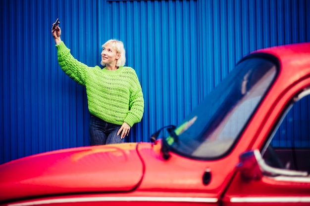 Selfie mobiele telefoon gelukkige mensen technologie concept met mooie blonde met groene kleding en rode vintage auto en blauwe muur. moderne millennial persoon trendy levensstijl en onafhankelijkheid