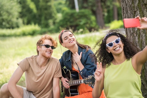 Selfie, picknick. Vrolijke ontspannen vrienden in zonnebril met gitaar die selfie op smartphone nemen bij picknick op zonnige dag