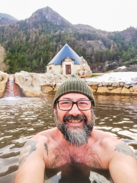 暖かい屋外の温泉水だけで楽しんでいる幸せな男の自撮り写真 雪と背景の山 冬と背景の山で自然の温泉水を楽しんでいる人々