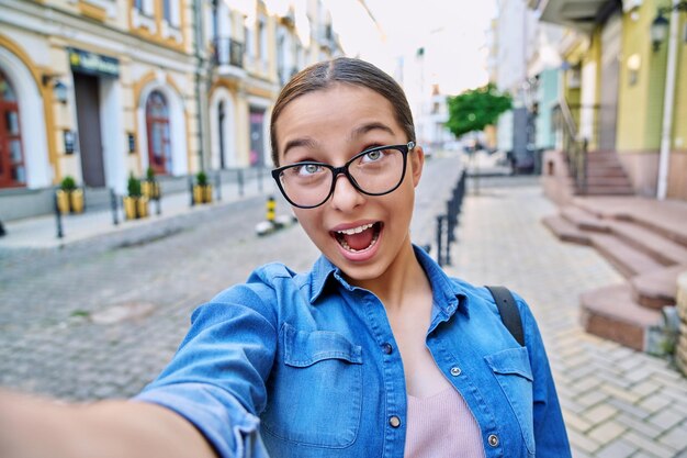 Selfie portrait of beautiful cheerful teenage girl outdoor on city street funny positive face close up Fun happiness emotions adolescence blog vlog youth concept