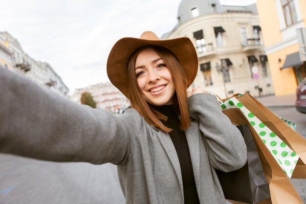 Фото Селфи-портрет. симпатичная привлекательная счастливая шопоголик женщина в осеннем пальто и шляпе, держащая много бумажных сумок для покупок в европейском городе