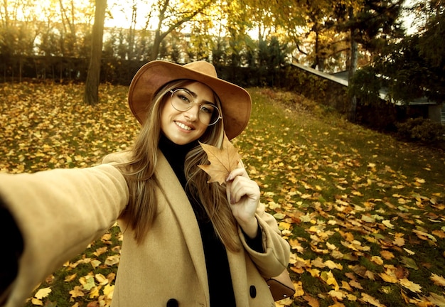 Selfie portrait of a funny young woman on the background of fallen leaves in the park. Autumn time