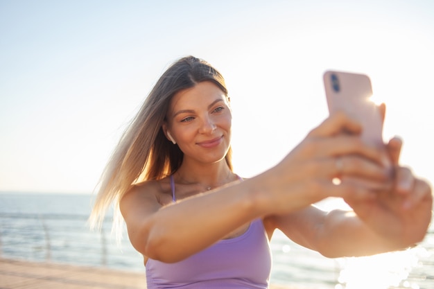 Selfie portrait of a young blonde woman dressed in sportswear on the beach at sunrise