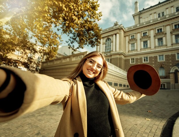 Selfie portrait of a young stylish tourist woman dressed in a coat and hat on a background of European urban architecture. Holidays and tourism concept