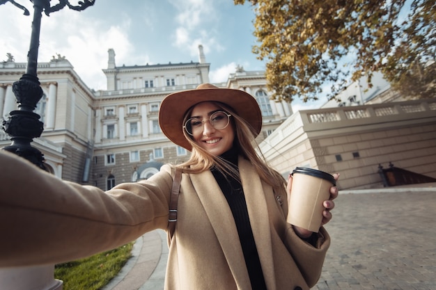 유럽 도시 건축의 배경에 코트와 모자를 입은 젊은 세련된 관광 여자의 셀카 초상화. 휴일 및 관광 개념