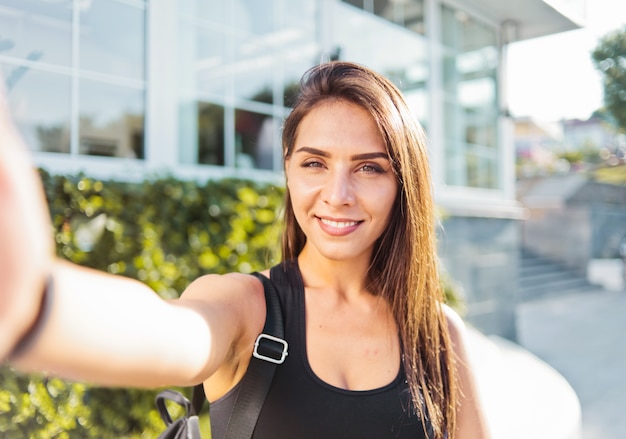 Selfie portret Jonge vrolijke fit vrouw in sport top met tas op haar schouders lachend buitenshuis