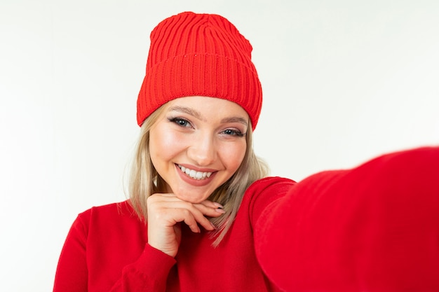 Selfie of a smiling blonde girl in a red hat and sweater on a white background