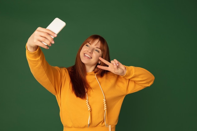 Selfie, sorridente. ritratto di donna caucasica isolato su sfondo verde studio con copyspace. bellissimo modello femminile con telefono. concetto di emozioni umane, espressione facciale, vendite, pubblicità, moda.