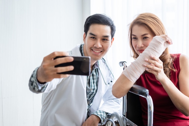 Selfie di un medico sorridente con una stecca per il braccio del paziente femminile per una migliore guarigione sedersi su una sedia a rotelle utilizzando uno smartphone sullo sfondo dell'ospedale della stanza.