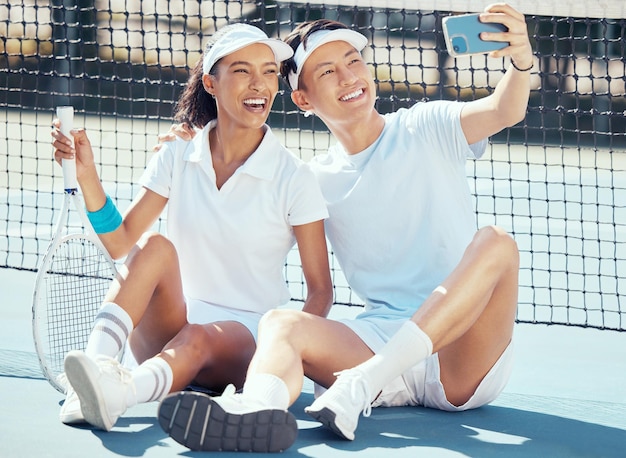 Selfie sports or couple sitting on a tennis court resting after exercise practice or training Wellness fitness or health with an interracial man and woman with sport racket taking a phone picture