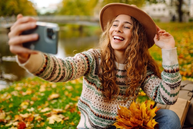 Selfie time Beautiful woman takes selfie on smartphone in hat on meadow in autumn park on plaid