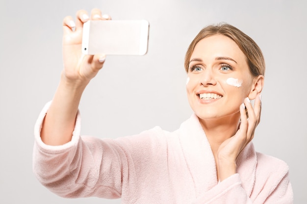 Foto tempo di selfie! ritratto di giovane bella donna sorridente mentre si prende un po 'di crema per il viso isolato su sfondo bianco con copia spazio.