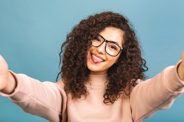 Selfie time. Young cheerful attractivecurly brunette is smiling isolated on blue background. She is taking selfie on the camera of her phone.