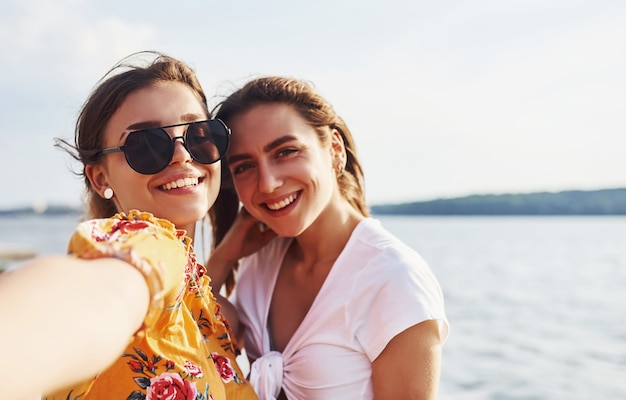 Photo selfie of two smiling girls outdoors that have a good weekend together at sunny day against the lake.