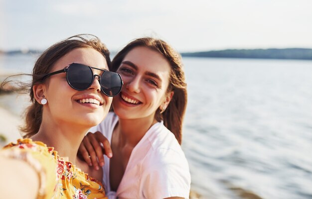 Selfie of two smiling girls outdoors that have a good weekend together at sunny day against the lake.