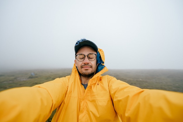 Selfie van een man met ogen gesloten in gele regenjas op verlaten plek met wolken en mist op de achtergrond.