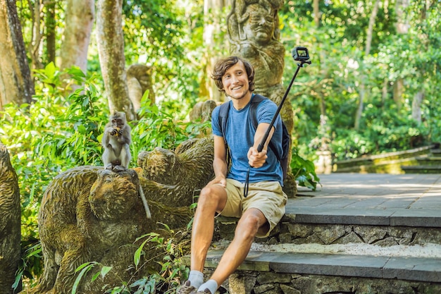 원숭이와 셀카. 젊은 남자는 셀카봉을 사용하여 귀엽고 재미있는 원숭이와 함께 사진이나 비디오 블로그를 찍습니다. 발리에서 야생 동물과 셀카를 여행하세요.