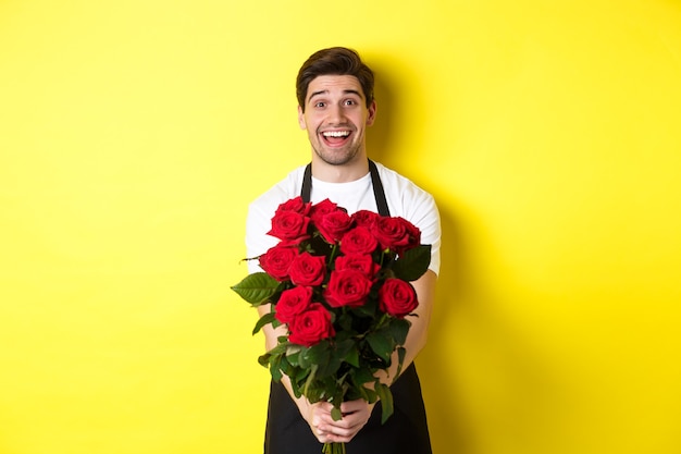 Seller in flower shop wearing black apron, giving bouquet of roses and smiling, standing over yellow wall