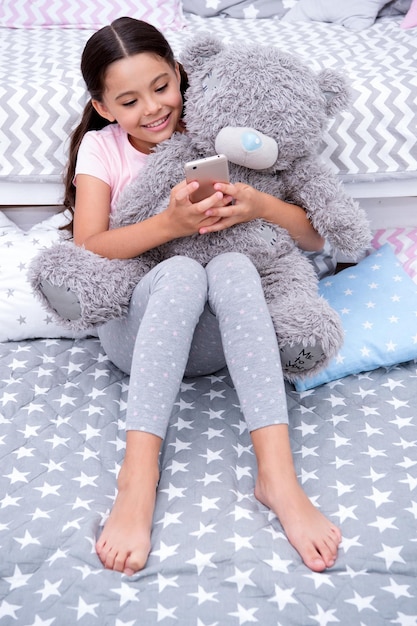 Send message sweet dreams Girl child sit on bed with teddy bear in her bedroom Kid prepare to go to bed Girl kid long hair cute pajamas relax plush teddy bear toy Child smartphone send message