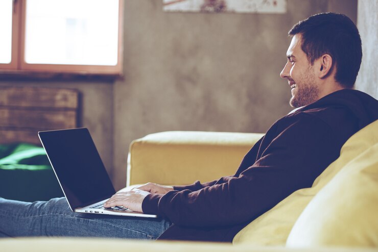 Photo sending e-mail. side view of cheerful young man using his laptop with smile while sitting on couch at home
