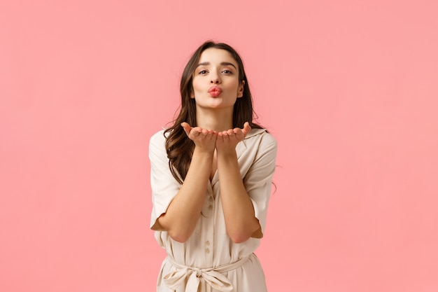 Sending love and care. Charming cute and tender european brunette with curly hair, wearing spring dress, leaning with hands near folded lips, blowing air kiss with love, standing pink wall