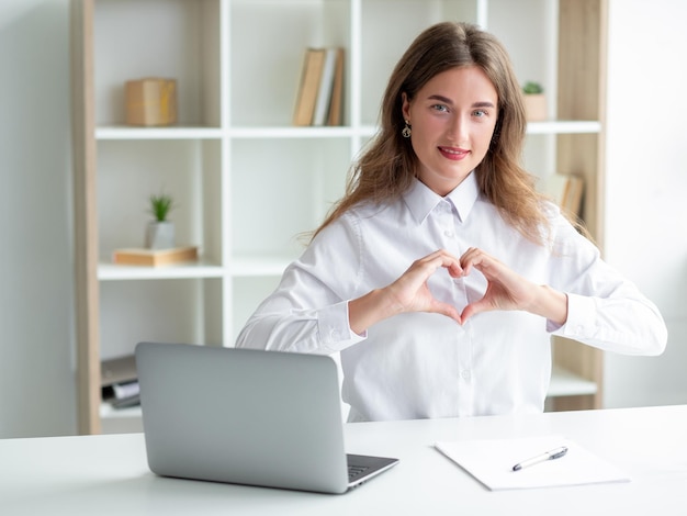 Sending love Office woman Inspired work Pretty smiling lady in white shirt sitting desk with laptop showing heart shape figure hands in light room interior