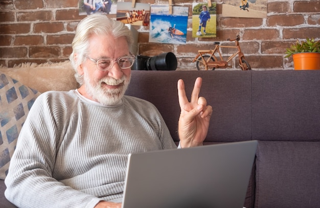 Senior aantrekkelijke man met laptop in videogesprek. oude gepensioneerde man die zijn tijd doorbrengt door thuis op een laptop te werken met camera, decoraties, foto's. bakstenen muur achtergrond