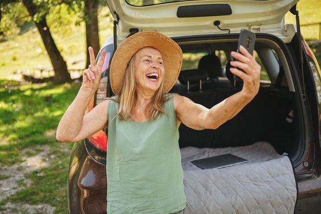 Senior active happy woman makes selfie with phone in car trunk or boot in camper sunny summer park