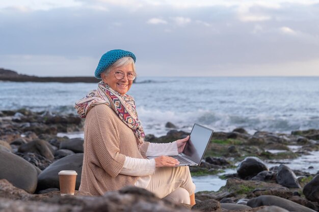 Senior active woman sitting on the pebble beach using laptop in remote work Mature female enjoying sunset at sea holding laptop on knees horizon over water