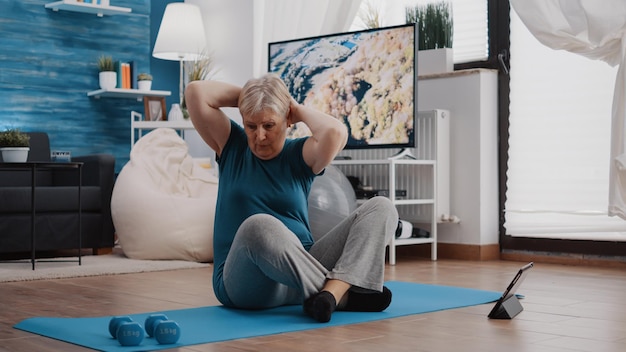 Photo senior adult doing physical exercise and watching video on tablet to follow online training lesson. old woman using device to do fitness workout and gymnastics from home. active person