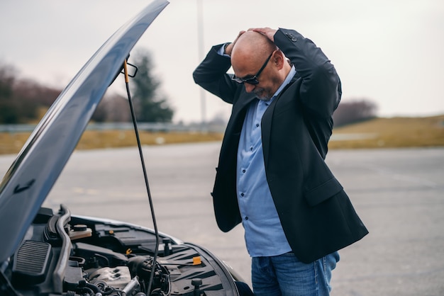 Senior adult man holding hands on head while standing in front of opened hood of his car.