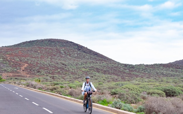 Senior adult woman wearing helmet and eyeglasses cycling in outdoors with backpack on shoulders Cloudy sky and mountain in background